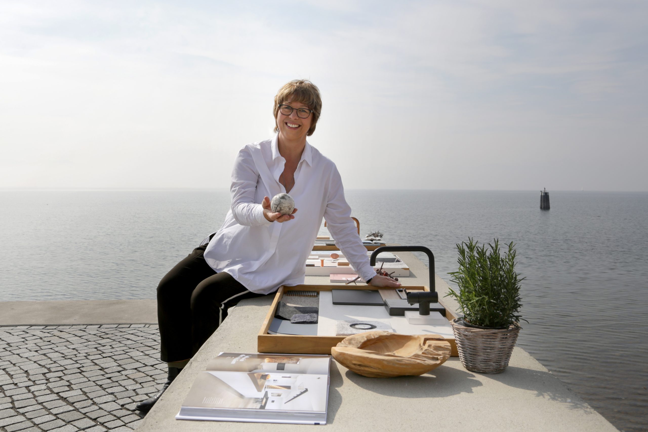 Frau mit Brille sitzt lächelnd auf Terrasse mit Kaffee, Laptop und Topfpflanze am Meer.