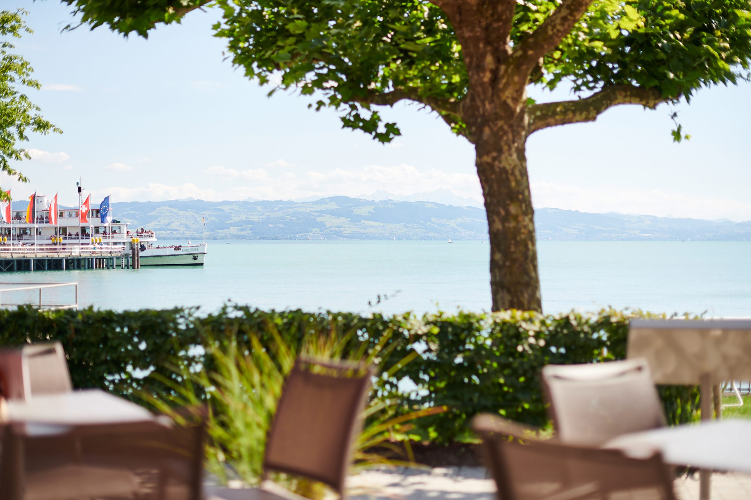 Blick vom Seehotel auf Steg mit Flaggen und See mit Bergen im Hintergrund