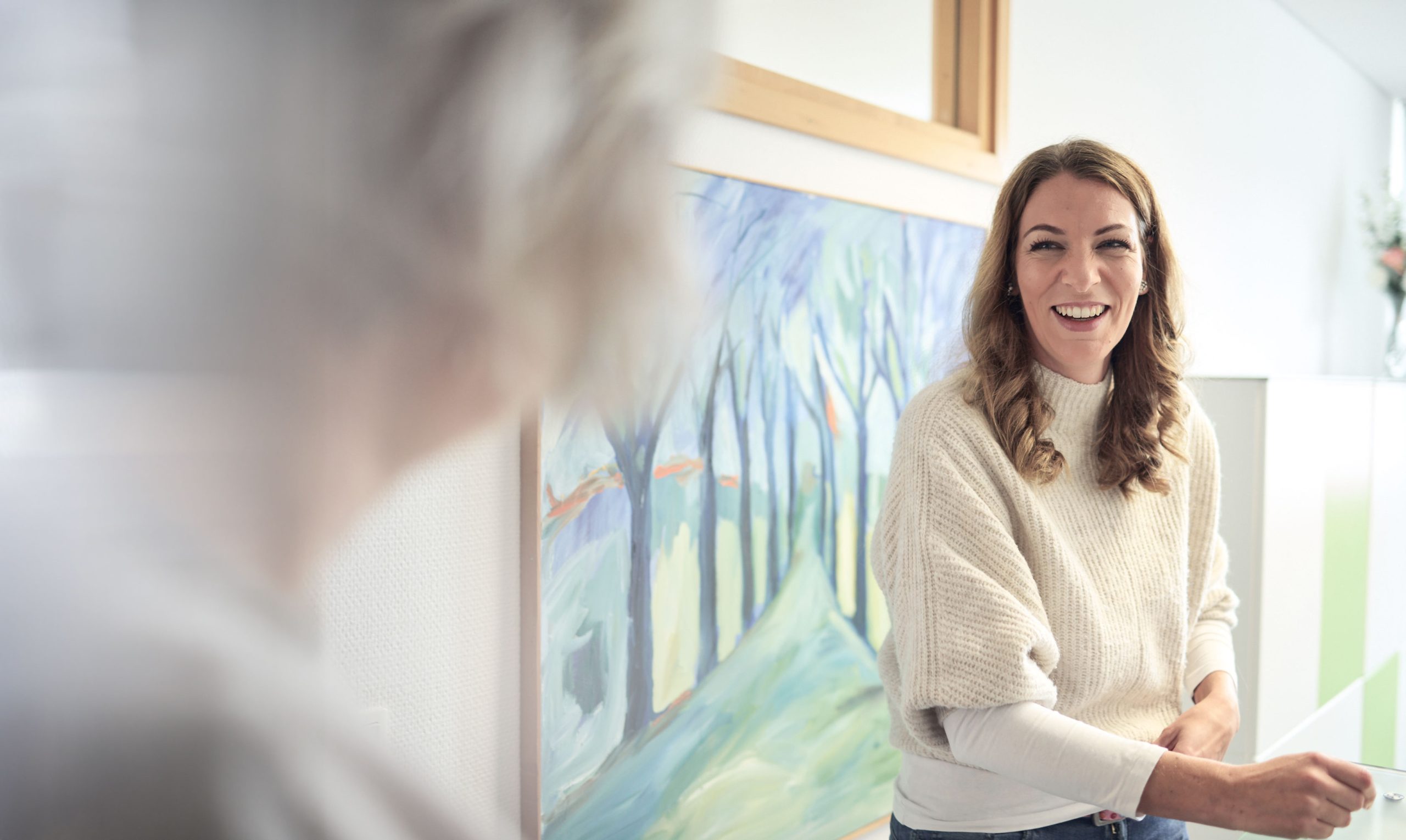 Frau mit langen braunen Haaren in beige-grauem Pullover vor hellblauem Vorhang.
